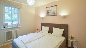 A double bed with white bedding, a window with blinds, a radiator, and a wall-mounted picture. - Comfortplus vacation apartment with 1 bedroom and terrace on the first floor (Ostseebad Binz)