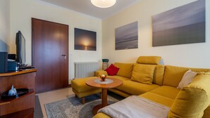 A living room with a yellow sofa, a small round table, a wooden cabinet, and a television. There are two paintings on the wall and a door to the left. - Komfort Ferienwohnung mit 2 Schlafzimmern und Balkon im Dachgeschoss (Ostseebad Binz)