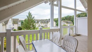 Ein überdachter Balkon mit weißen Stühlen und einem Tisch, mit Blick auf eine Wohngegend mit Häusern und Grünflächen.