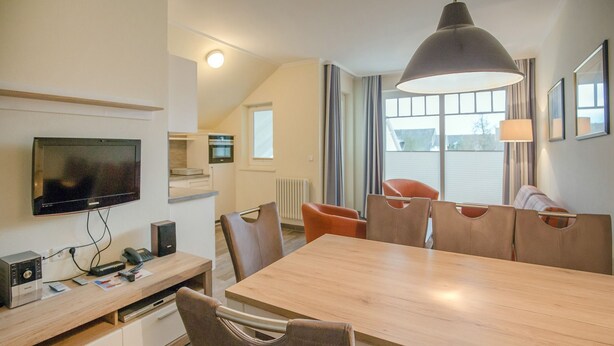 Ein modernes Hotelzimmer mit Essbereich, einem Flachbildfernseher, einem Schreibtisch mit Computer und einem großen Fenster mit Vorhängen.