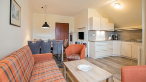 Ein modernes Wohnzimmer mit Sofa, Sesseln und einem Esstisch. Es gibt eine Küchenzeile mit Schränken und einer Arbeitsfläche.
