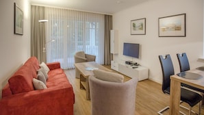 Un salon moderne avec un canapé rouge, un fauteuil beige et une table basse en bois. Un téléviseur à écran plat est fixé au mur, il y a une table à manger avec des chaises, et une grande fenêtre ornée de rideaux légers.