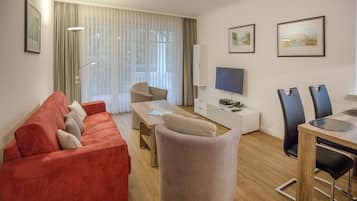 A modern living room with a red sofa, a beige armchair, and a wooden coffee table. There is a flat-screen TV mounted on the wall, a dining table with chairs, and a large window with sheer curtains.