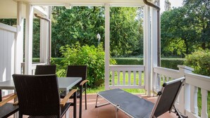 Une terrasse couverte avec une table et des chaises, un banc et une vue sur un jardin agrémenté d’arbres et d’une haie.