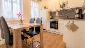 Une cuisine moderne avec des meubles blancs, un four encastré, un four à micro-ondes et une machine à café. On y trouve une table à manger avec des chaises et un vase rempli de fleurs.