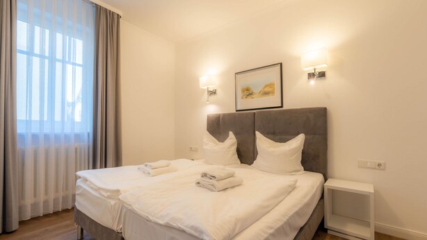 Un lit double avec du linge de lit blanc, une tête de lit, des lampes de chevet et un tableau encadré au mur.