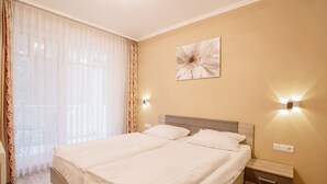 Uma cama de casal com lençóis brancos, cabeceira de madeira, duas mesas de cabeceira com gavetas, luminária fixada na parede e uma janela grande com cortinas leves.