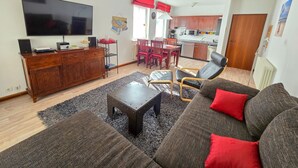 Una sala de estar con un sofá, una mesa de centro y un televisor. Se observa una cocina al fondo con una mesa de comedor y sillas.