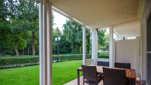 Un patio techado con mesa y sillas, con vistas a un jardín y árboles bien cuidados.