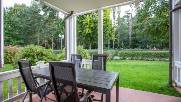 Un porche cubierto con una mesa y sillas, con vistas a un jardín bien cuidado con setos y árboles.