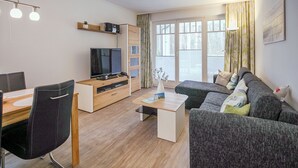 Una sala de estar moderna con un sofá, un televisor sobre un mueble de madera, una mesa de comedor con sillas y ventanas amplias con cortinas.