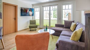 Una sala de estar con un sofá, un sillón y una pequeña mesa. Se ve una puerta y una ventana con vistas al exterior.