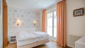 Un dormitorio con cama, mesita de noche, lámpara de pared, ventana con cortinas y radiador.