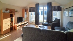 Ein modernes Wohnzimmer mit Flachbildfernseher, Holzmöbeln, einer Couch und einem Balkon mit Vorhängen.