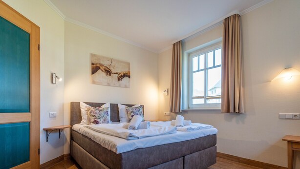A modern bedroom with a large bed, a small bedside table, a wall-mounted light, and a window with curtains. - Comfort apartment with 2 bedrooms and terrace on the first floor (Ostseebad Binz)