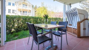 A covered patio with a table and chairs, a wicker chair, and a view of a building and greenery. - Comfort apartment with 2 bedrooms and terrace on the first floor (Ostseebad Binz)