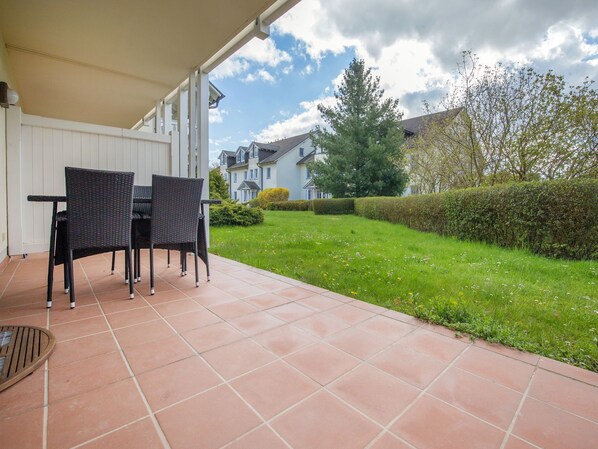 A patio with two wicker chairs and a table, a tiled floor, and a view of a well-maintained lawn and neighboring houses.