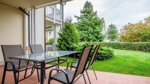 An outdoor patio with a glass table and four chairs, a tiled floor, and a view of a green lawn and trees.