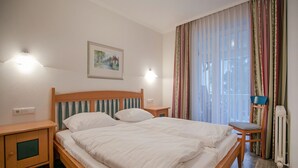 Un lit bien fait, avec une literie blanche, une tête de lit en bois et deux tables de chevet avec des lampes. Un tableau encadré est accroché au mur au-dessus du lit.