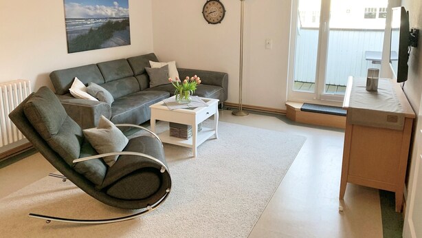 A modern living room with a grey sofa, a rocking chair, a coffee table with a vase of flowers, a floor lamp, and a large window with a view of the outdoors. - Comfort apartment with 2 bedrooms and balcony on the top floor (Ostseebad Binz)