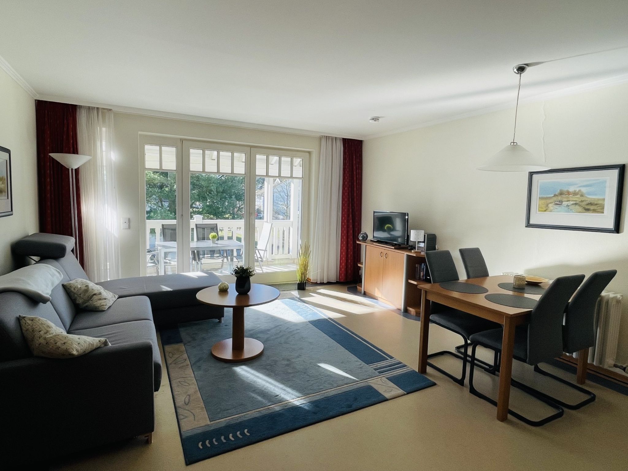 A modern living room with a sofa, a dining table with chairs, a TV, and a view of the outdoors through large windows.