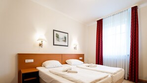 Cama doble con ropa blanca, cabecera de madera, mesitas de noche, lámparas de pared, un cuadro enmarco y cortinas rojas.