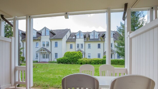 Une terrasse couverte avec deux chaises blanches et une table, donnant sur une pelouse bien entretenue et une rangée de maisons.