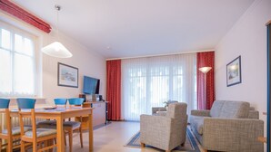 Ein Wohnzimmer mit Essbereich, zwei Sessel, einer Couch, einem Fernseher und einem Fenster mit Vorhängen.