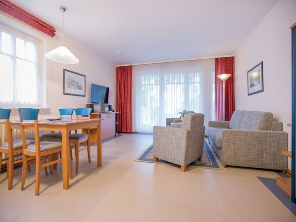 Ein Wohnzimmer mit Essbereich, zwei Sessel, einer Couch, einem Fernseher und einem Fenster mit Vorhängen.