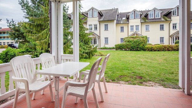 Eine überdachte Terrasse mit weißen Plastikstühlen und einem Tisch, mit Blick auf einen gepflegten Rasen und eine Reihe von Häusern.