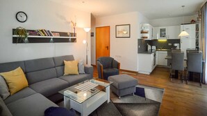 Een moderne woonkamer met een bank, voetenbankje en salontafel, een kleine eethoek met stoelen en een kitchenette met witte kasten en een spoelbak.