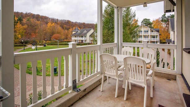 Een overdekt terras met witte leuningen, een tafel met stoelen en uitzicht op een woonwijk met herfstkleurige bomen.