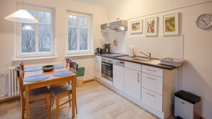A modern kitchen with white cabinets, a dishwasher, a stove, and a sink. There are two windows providing natural light and a clock on the wall.