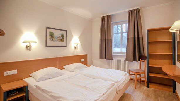 A hotel room with a double bed, two bedside tables, a chair, a desk, and a window with curtains.