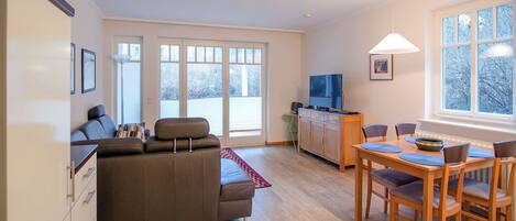 A modern living room with a sofa, dining table, and chairs. There is a television on a cabinet and a window with a view of trees.