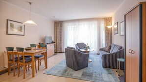 A modern living room with a dining area, a sofa set, a TV, and a large window with curtains. - Comfort apartment with 1 bedroom and terrace on the first floor (Ostseebad Binz)