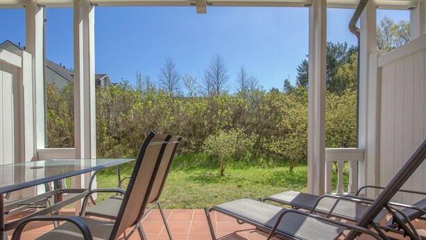 A covered patio with a table and chairs, a view of a garden with trees and shrubs, and a clear blue sky. - Comfort apartment with 1 bedroom and terrace on the first floor (Ostseebad Binz)