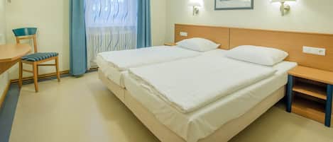 A hotel room with a double bed, a desk with a chair, a nightstand, and a window with curtains.