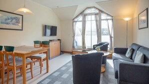 A cozy living room with a dining area, a flat-screen TV mounted on the wall, and a large window with curtains. - Budget Ferienwohnung mit 1 Schlafzimmer und Balkon im Dachgeschoss (Ostseebad Binz)