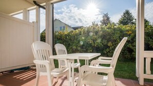 Eine Terrasse im Freien mit einem weißen Tisch und vier weißen Stühlen, umgeben von einem Garten und einem Haus im Hintergrund.