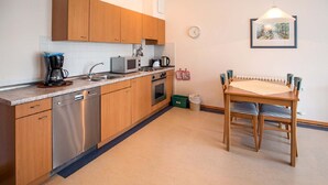 A compact kitchen with wooden cabinets, a dishwasher, a microwave, and a coffee maker. There's a dining table with chairs and a hanging light fixture.