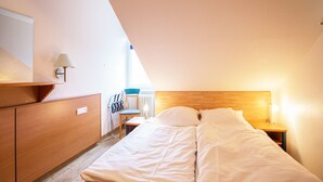 A bedroom with a wooden headboard, a bed with white bedding, a nightstand with a lamp, and a chair with a blue cushion. - Comfort apartment with 1 bedroom and balcony on the top floor (Ostseebad Binz)