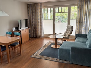 A room with a dining table, chairs, a sofa, a TV, and a window with curtains.