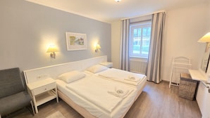 Ein Hotelzimmer mit einem großen Bett, Nachttischen, einem Schreibtisch, einem Sessel, einem Fenster mit Vorhängen und einem Bild an der Wand.