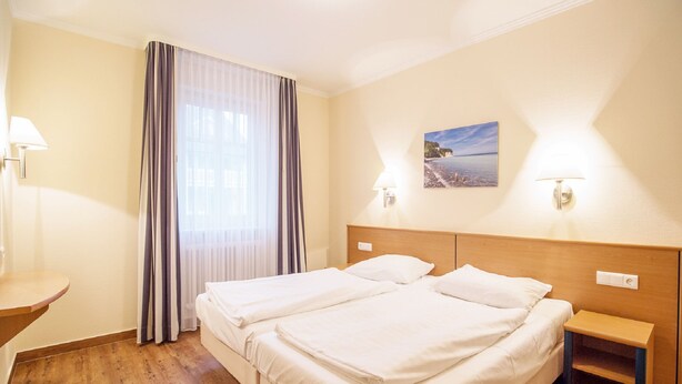Hotellihuone, jossa on parivuode, yöpöytä, seinävalaisin, ikkuna verhoilla ja seinään kiinnitetty lamppu.