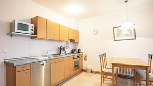 A compact kitchen with wooden cabinets, a built-in dishwasher, and a microwave. There's a dining table with chairs and a hanging light fixture. - Comfort apartment with 1 bedroom and balcony on the upper floor (Ostseebad Binz)