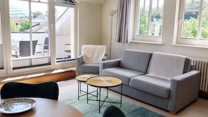 Una sala de estar con un sofá gris, una mesa redonda y una mesa de comedor con sillas. Hay ventanas grandes con cortinas, un radiador y vistas a casas.