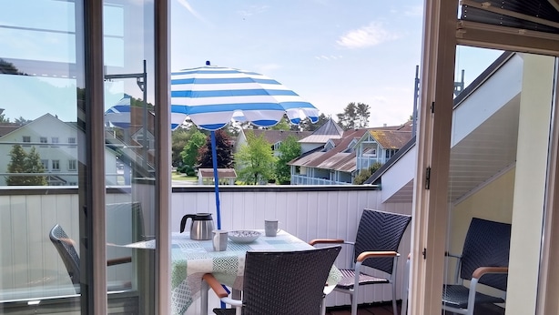 Un balcón con mesa y sillas, una sombrilla azul y blanca, y vistas a casas al fondo.