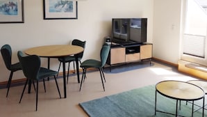 Una sala de estar moderna con una mesa de comedor redonda, dos sillas, un televisor sobre un mueble de madera y cuadros enmarcados en la pared.