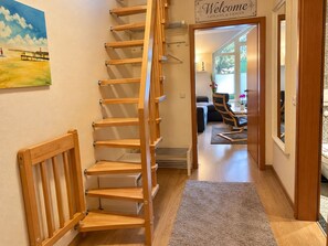 Un escalier en colimaçon en bois, surmonté d’une pancarte indiquant « Bienvenue à vos amis et à votre famille », mène à un espace de vie comprenant un canapé, un fauteuil et une table.